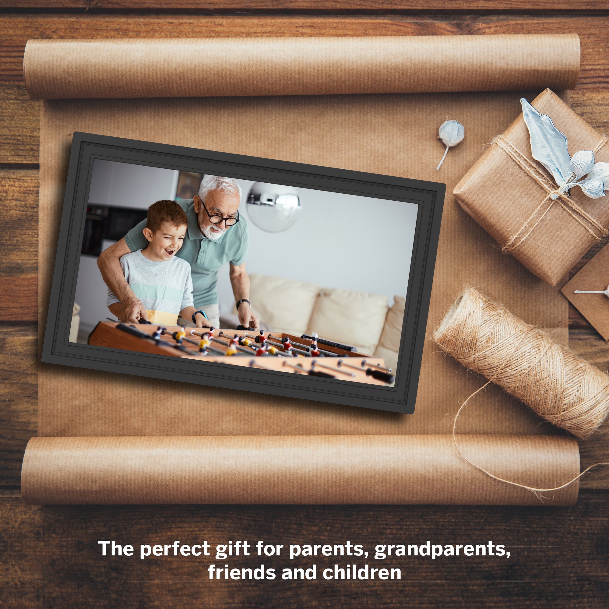 Digital photo frame showing an older man and a child playing with a foosball table, surrounded by gift wrapping materials on a wooden surface.
