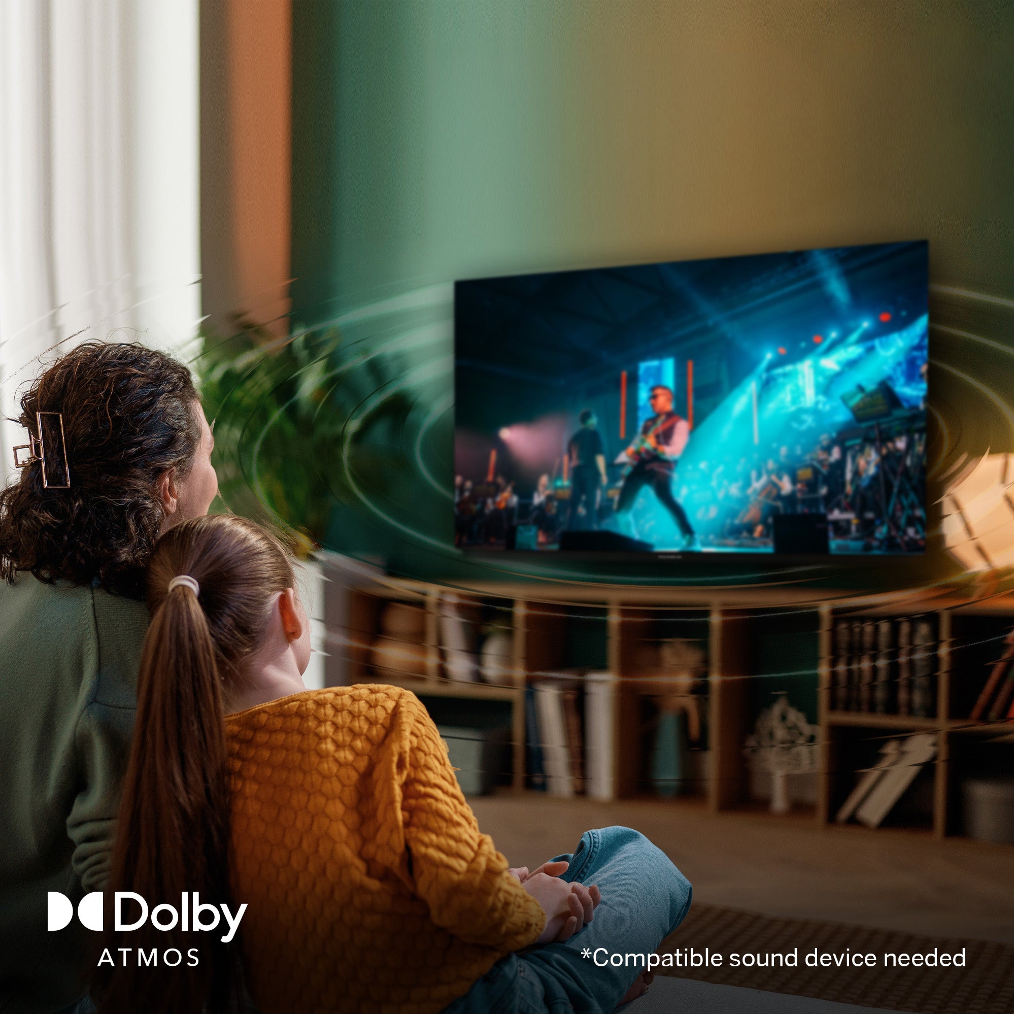 Two people sitting in a living room watching a concert on a large TV screen, with Dolby Atmos logo.