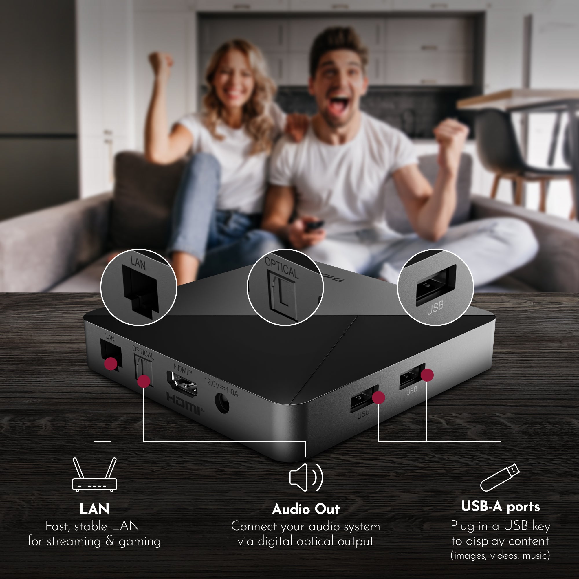 Black streaming device with ports labeled on a wooden surface, with a blurred background of two people sitting on a couch.